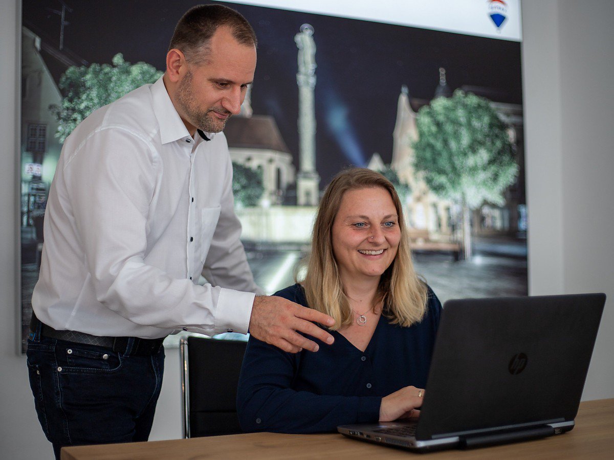 Immobilienmakler Schongau - Stockhausen Immobilienteam berät Sie kompetent beim Kauf, Verkauf und der Bewertung Ihrer Immobilie in Schongau in Oberbayern Stockhausen Immobilienmakler Schongau - zwei Makler arbeiten gemeinsam am Laptop im Büro vor Wandbild des Schongauer Marktplatzes